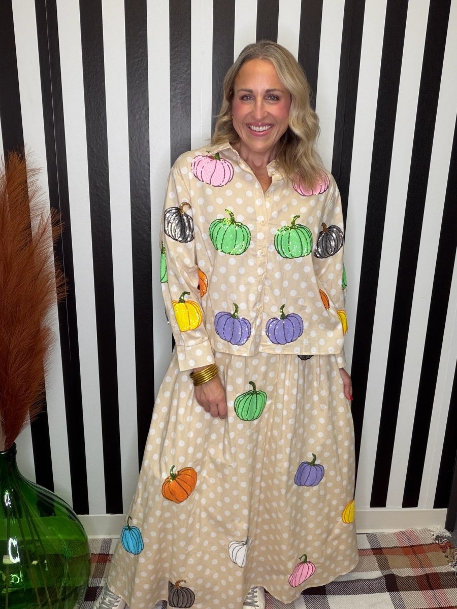 A woman wearing a matching long-sleeve button-up shirt and skirt set with a beige and white polka dot pattern and colorful pumpkin prints, standing in front of a black and white striped wall.