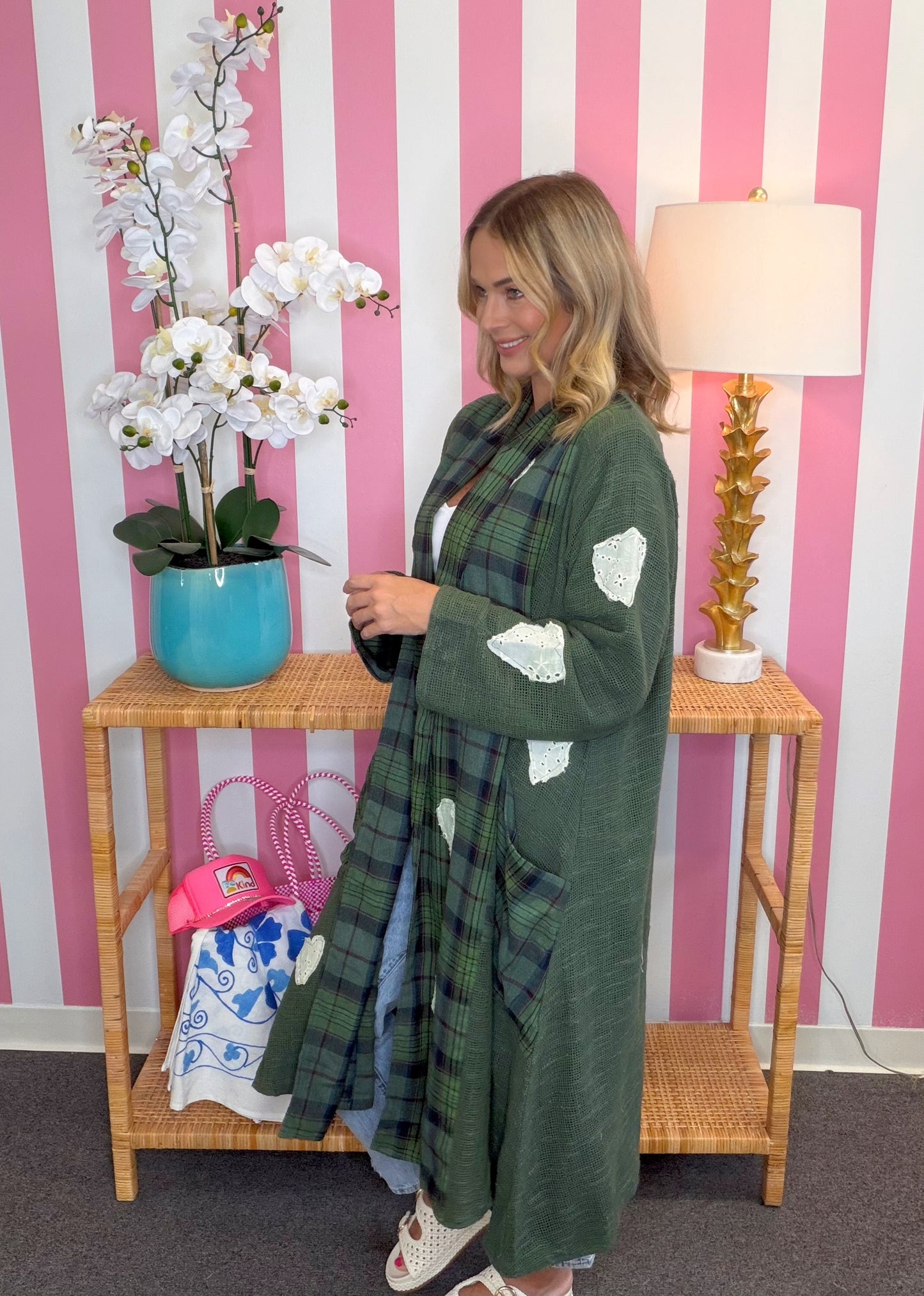 A woman wearing a long green plaid cardigan with white patchwork details stands in front of a pink and white striped wall, next to a wooden console table with a blue orchid pot and a lamp.