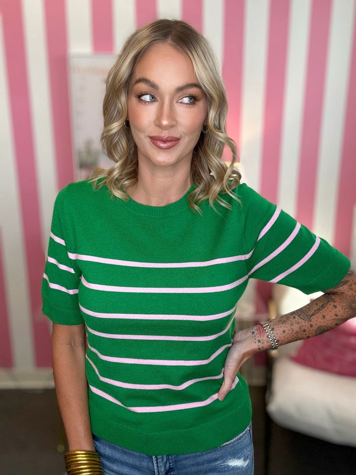 A woman wearing a green short-sleeve knit top with horizontal white stripes, paired with blue jeans, standing in front of a pink and white striped wall.