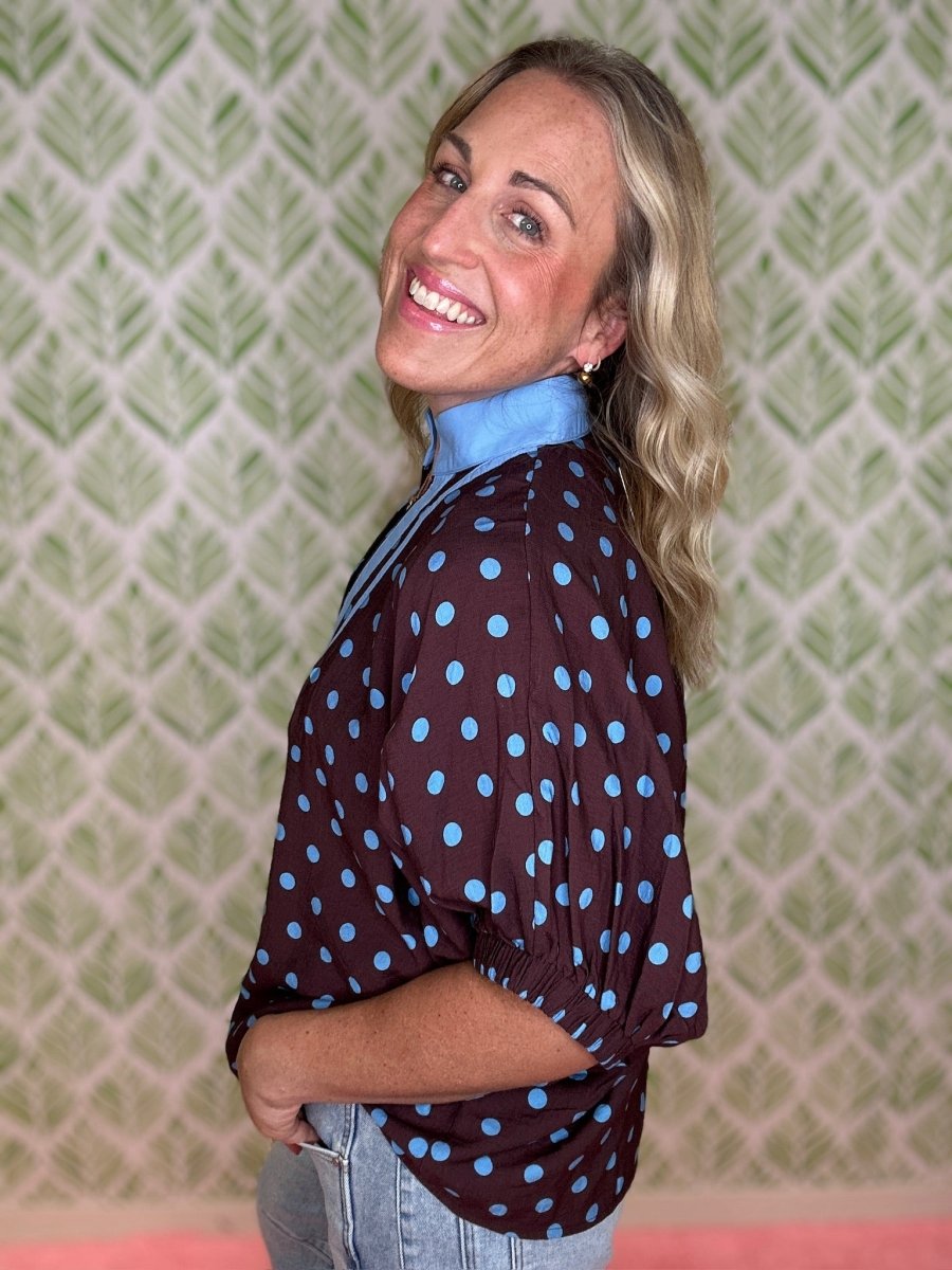 A smiling blonde woman wearing a dark brown long-sleeve blouse with light blue polka dots and a light blue collar, paired with light-wash jeans, standing in front of a green and white patterned wallpaper background.