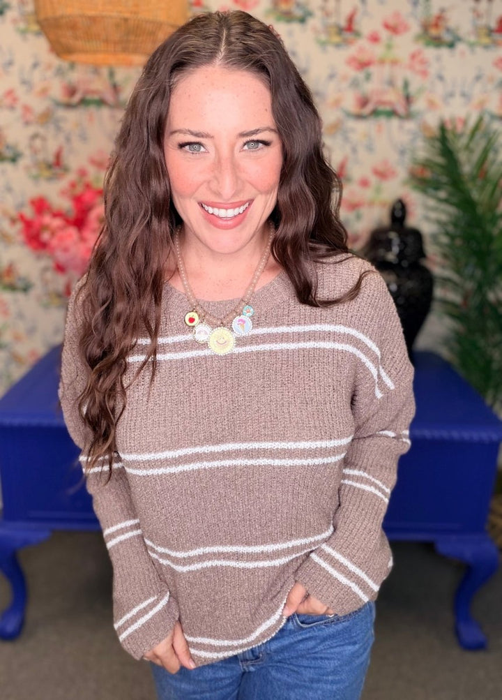 A smiling woman with long wavy brown hair wearing a brown knit sweater with white horizontal stripes and a colorful charm necklace, standing in front of a floral wallpaper background with a blue bench.
