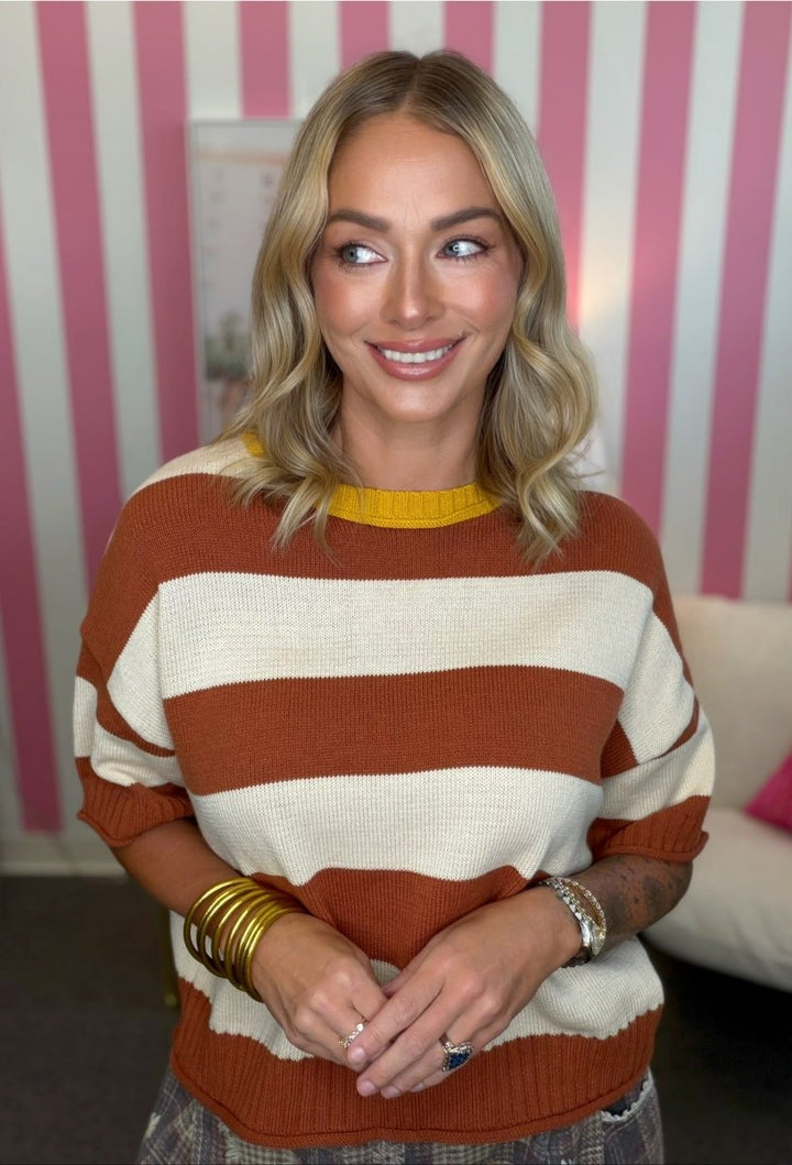 A woman with blonde hair wearing a brown and cream striped knit sweater with a yellow collar, standing in front of a pink and white striped wall.