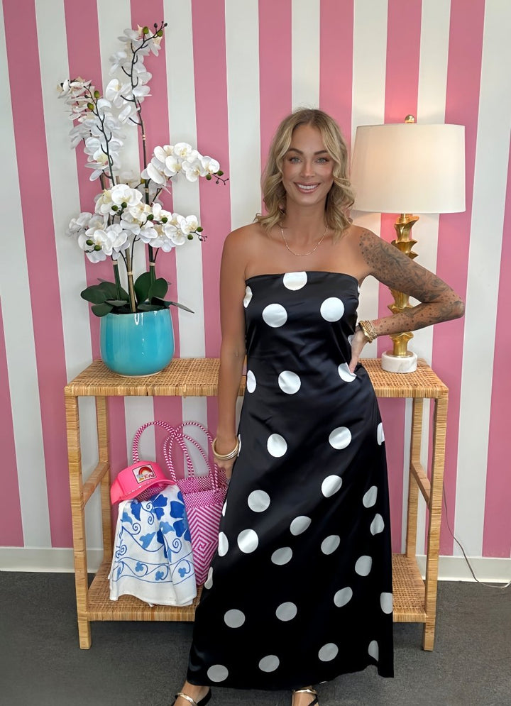 A woman modeling a strapless black satin maxi dress with large white polka dots, standing in front of a pink and white striped wall next to a wooden side table with a lamp and flowers.