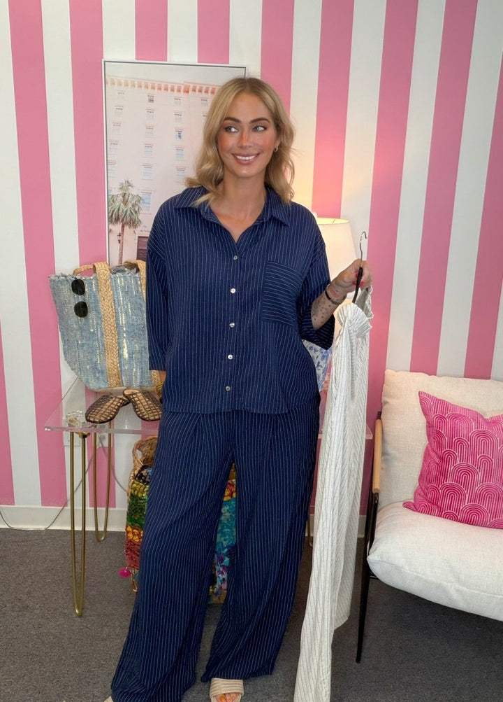 A woman stands indoors wearing a navy blue pinstriped button-up shirt and matching wide-leg pants, holding a white pinstriped garment on a hanger. The background features bold pink and white vertical stripes, with a side table, tote bag, and a white chair with a pink pillow visible.