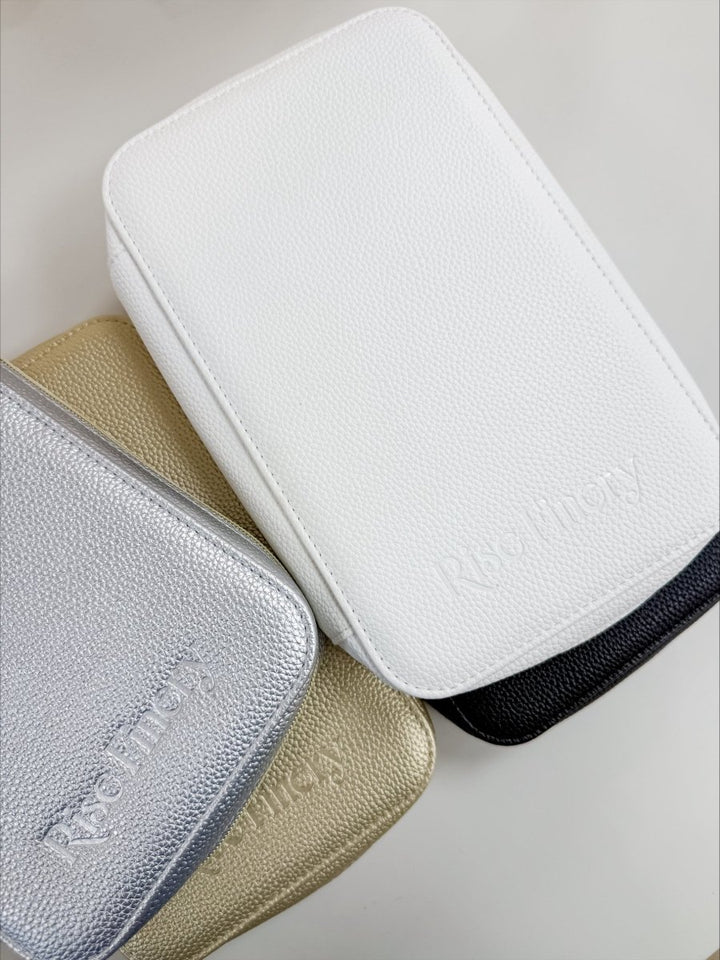 Three textured leather zippered pouches stacked diagonally on a light neutral background. The top pouch is white, the bottom-left is metallic silver, the bottom-center is metallic gold, and a black edge is visible behind the gold pouch.