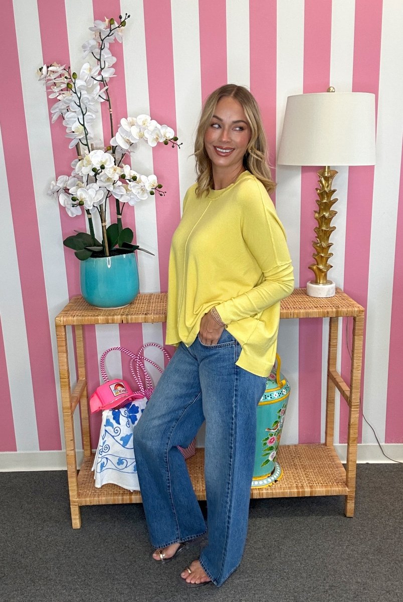 A woman stands in a room with pink and white striped wallpaper, wearing a bright yellow long-sleeve top and blue jeans. She is posed next to a wooden console table decorated with a potted white orchid, a turquoise vase, and a lamp.