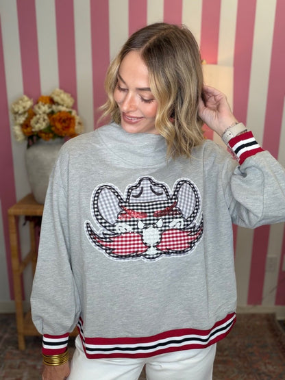 A woman wearing a light gray crewneck sweatshirt with a large embroidered gingham and red striped cow graphic on the front, paired with white pants, set against a pink and white striped wall background.