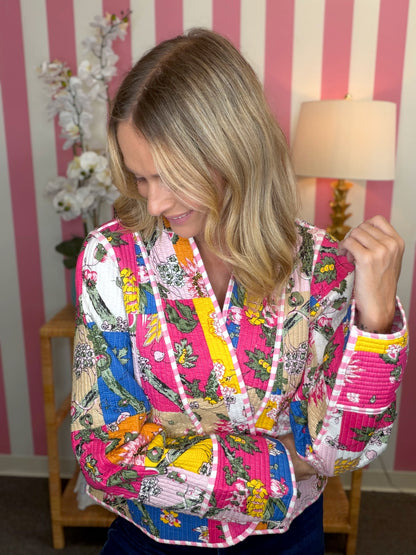 A woman wearing a vibrant, multicolor patchwork quilted jacket with floral and botanical patterns, standing in front of a pink and white striped wall.