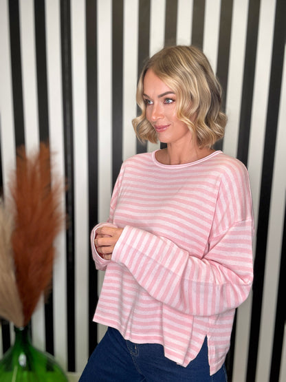A woman with blonde wavy hair wearing a pink and white striped long-sleeve top and dark blue jeans, standing in front of a black and white striped wall.