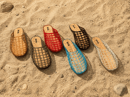 Five woven sandals in various colors on a sandy surface