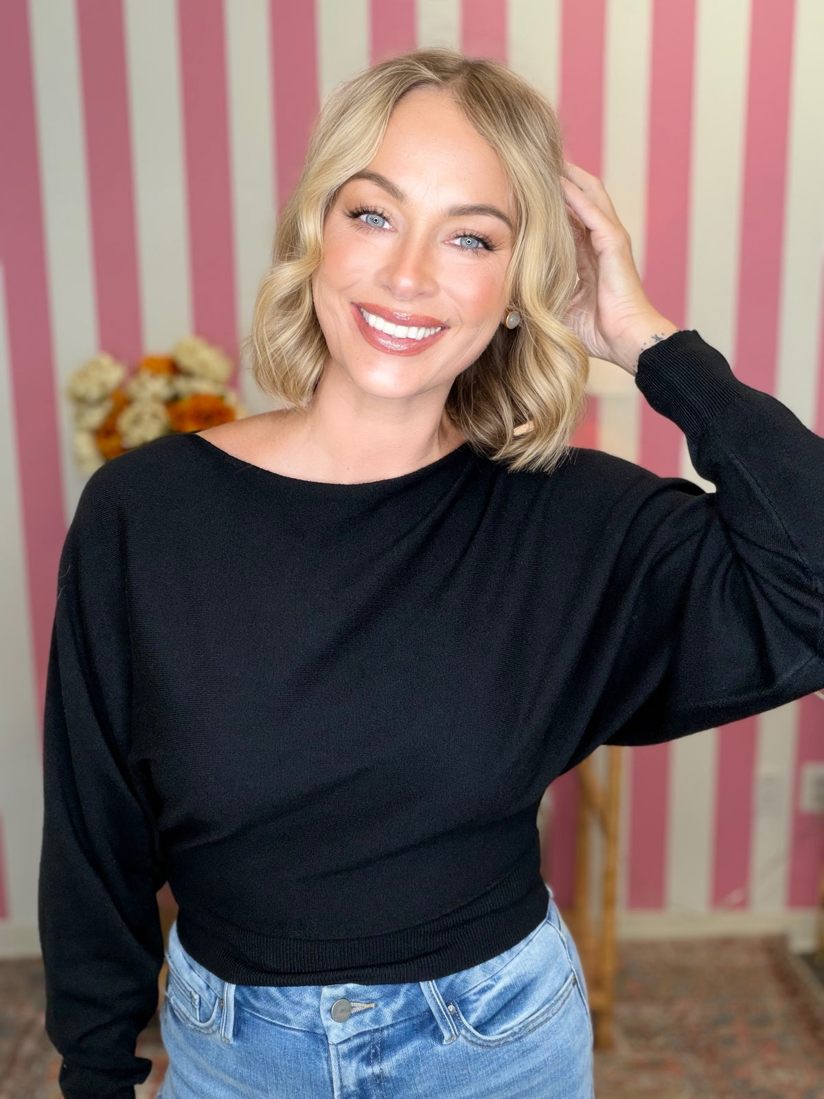 A smiling blonde woman wearing a black long-sleeve boat-neck top and light blue jeans, posing in front of a pink and white striped wall.