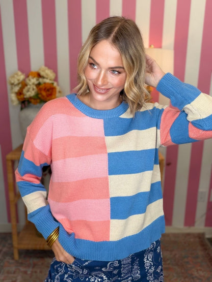 A woman wearing a colorful knit sweater with large pink, blue, and cream horizontal stripes, standing in front of a pink and white striped wall.