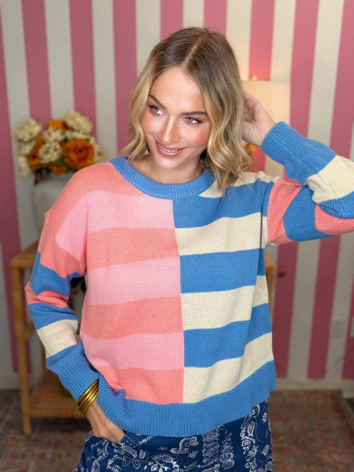 A woman wearing a colorful knit sweater with large pink, blue, and cream horizontal stripes, standing in front of a pink and white striped wall.
