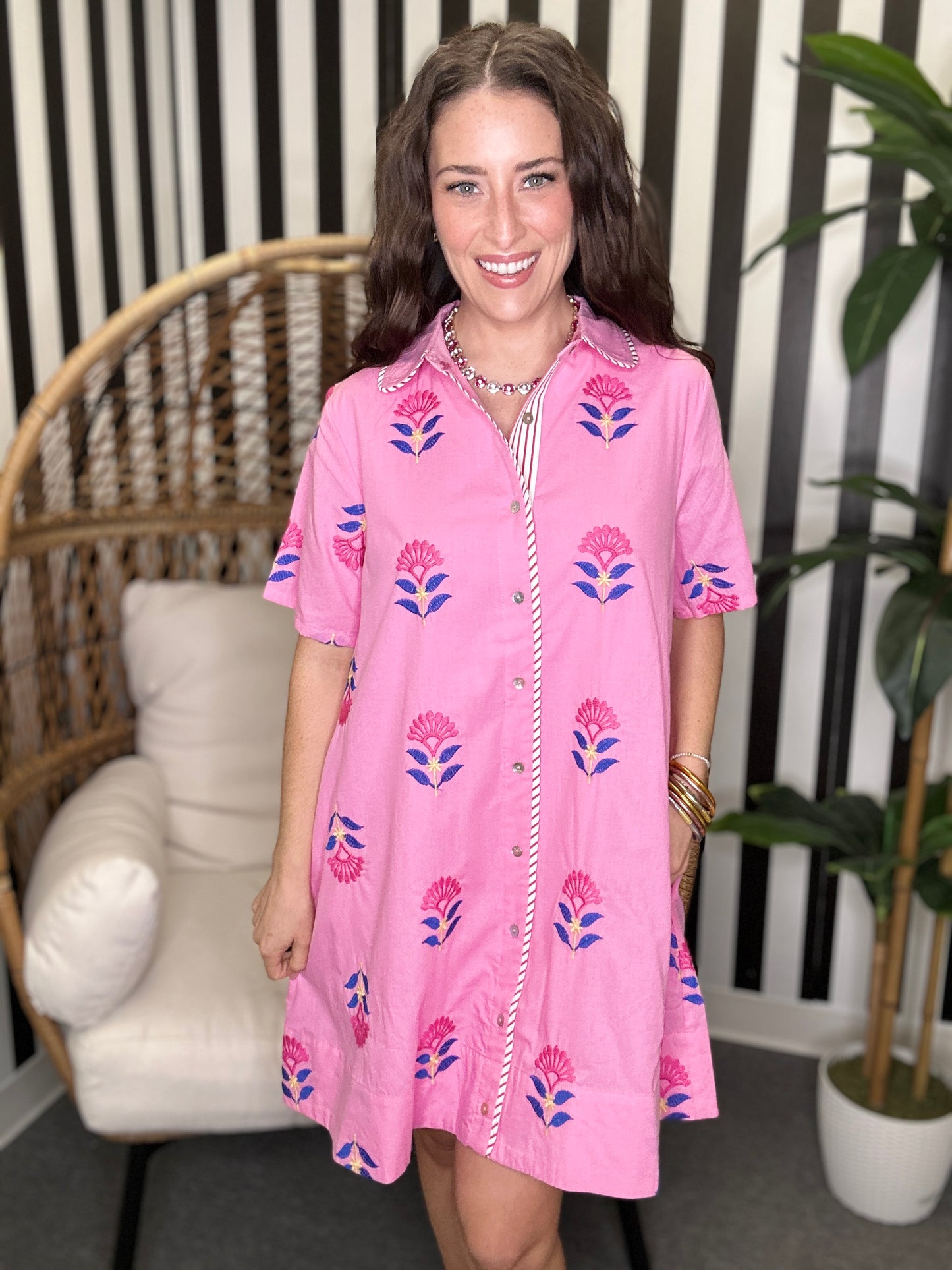 Woman wearing a pink floral dress standing in a room with a chair and plant.