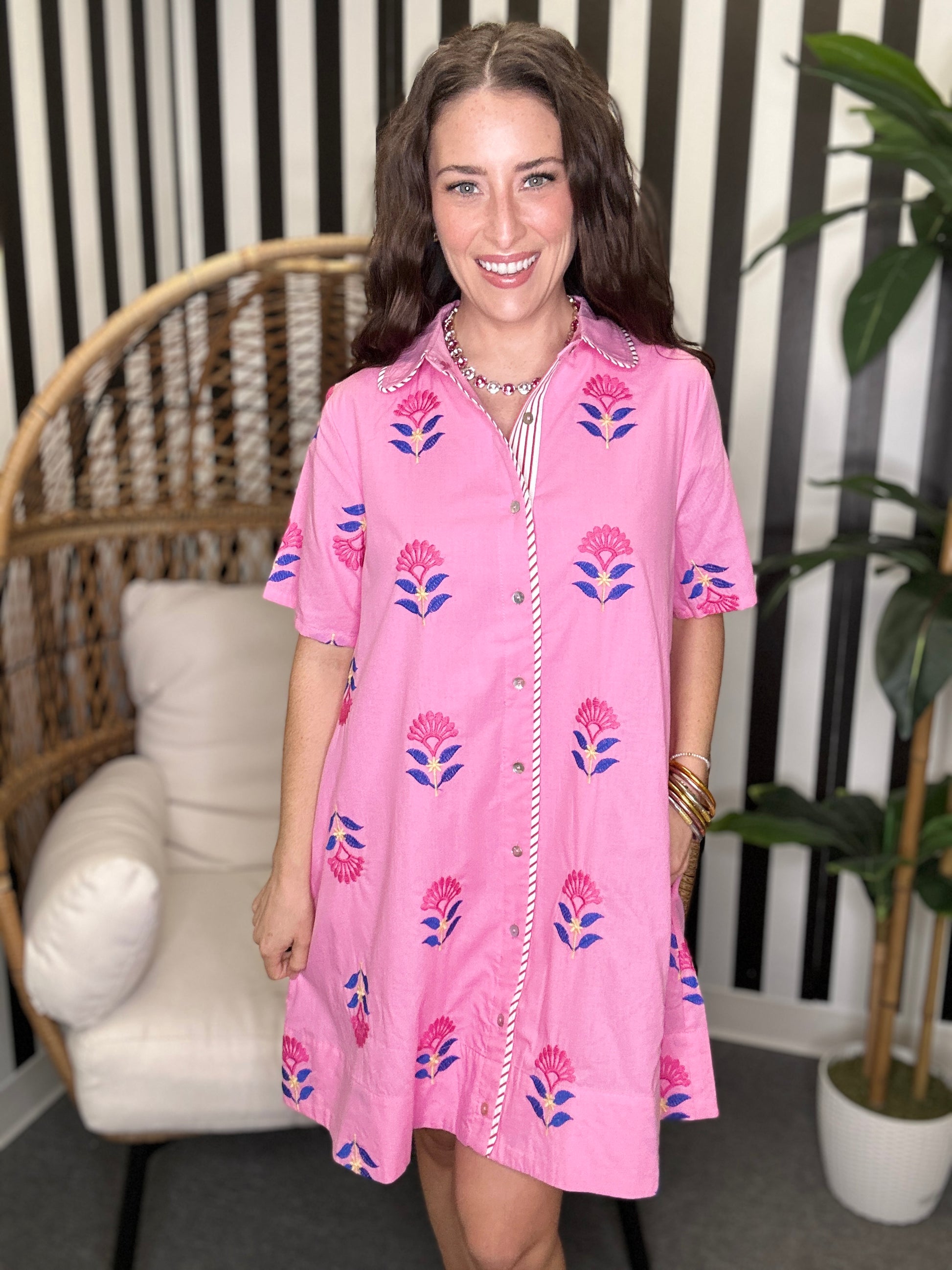 Woman wearing a pink floral dress standing in a room with a chair and plant.
