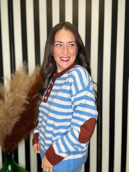 A woman wearing a blue and white striped knit sweater with brown trim and elbow patches, standing in front of a black and white striped wall.