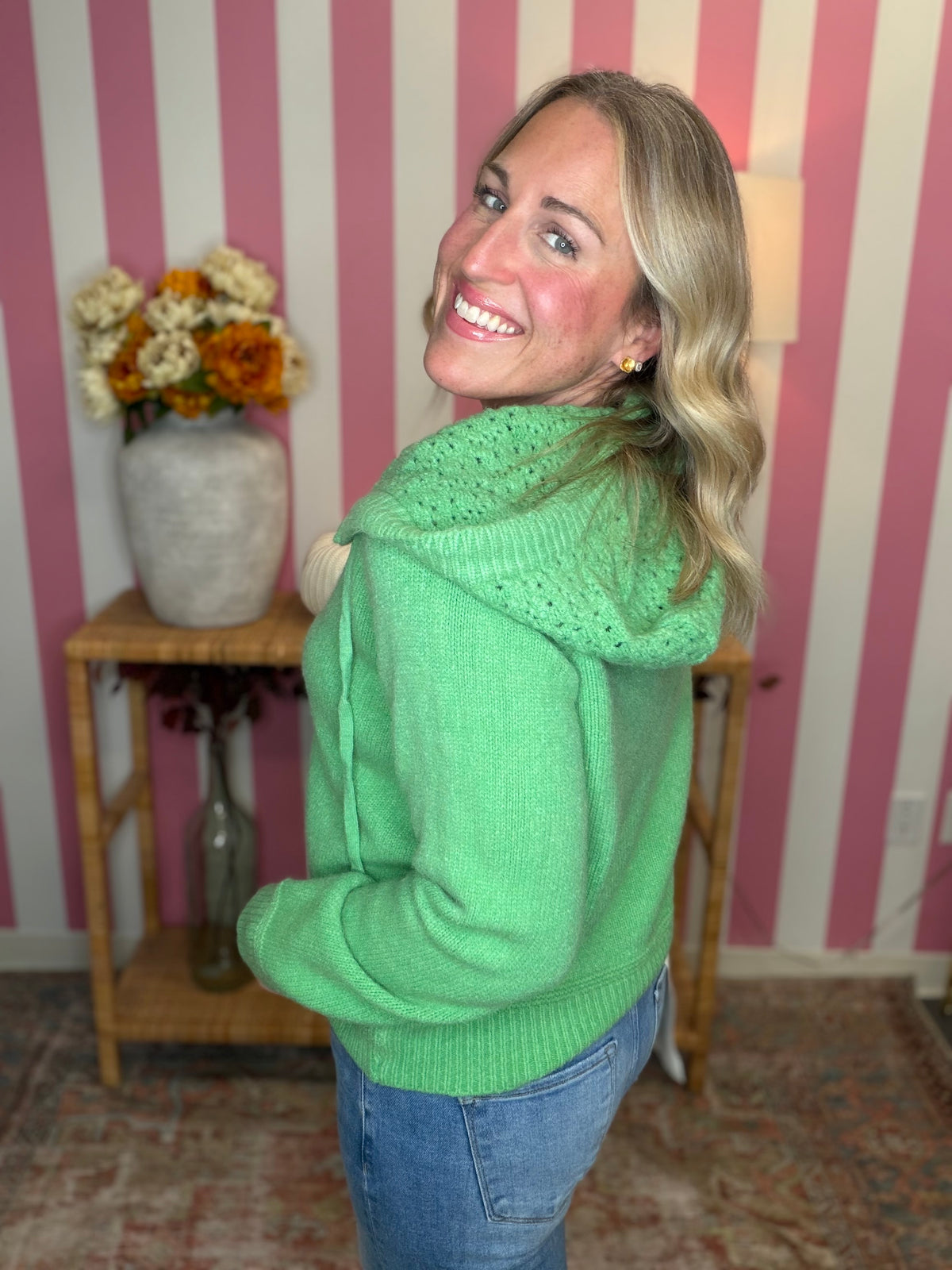 A smiling blonde woman wearing a bright green knit hooded sweater and blue jeans, standing in front of pink and white striped wallpaper with a wooden side table and vase of flowers in the background.