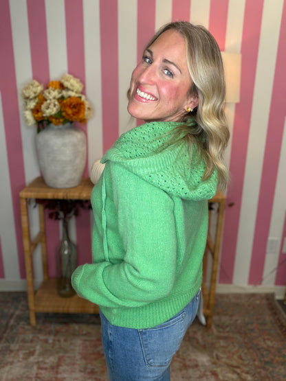 A smiling blonde woman wearing a bright green knit hooded sweater and blue jeans, standing in front of pink and white striped wallpaper with a wooden side table and vase of flowers in the background.