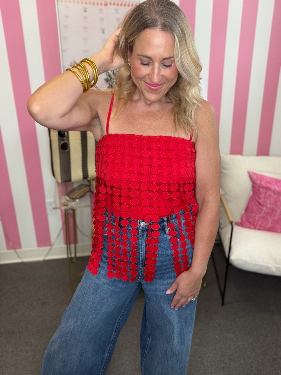 A woman wearing a bright red spaghetti-strap top with a textured circular pattern and fringe, paired with blue jeans, standing in front of a pink and white striped wall.