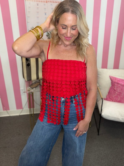 A woman wearing a bright red spaghetti-strap top with a textured circular pattern and fringe, paired with blue jeans, standing in front of a pink and white striped wall.