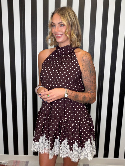 A woman modeling a sleeveless halter-neck mini dress with a dark brown and white polka dot pattern and a white lace hem, standing in front of a black and white striped wall.