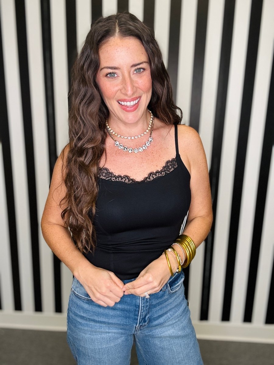 A woman with long wavy brown hair smiling, wearing a black lace-trimmed camisole top and blue jeans, accessorized with a silver necklace and gold bracelets, standing in front of a black and white striped wall.