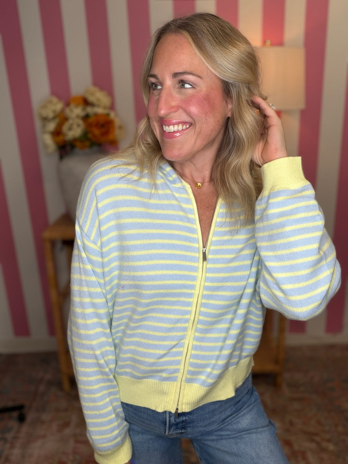 A smiling blonde woman wearing a pastel yellow and light blue striped zip-up sweater with yellow ribbed cuffs and hem, paired with blue jeans, standing in front of a pink and white striped wall.