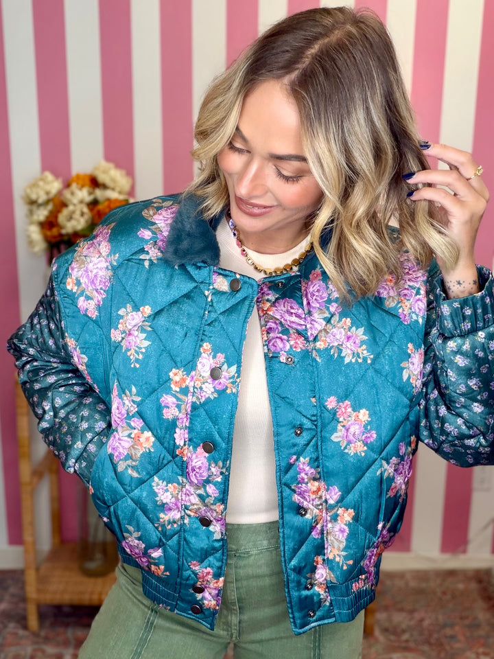 A woman wearing a teal floral quilted bomber jacket with a fuzzy collar, styled with olive green pants and a white top, standing in front of a pink and white striped wall.