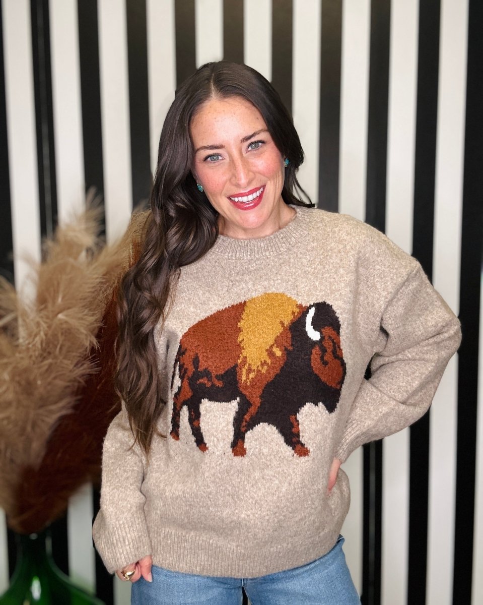 A woman smiling while wearing a beige knit sweater with a large bison graphic in brown, black, and tan on the front, paired with blue jeans, standing against a black and white striped background.