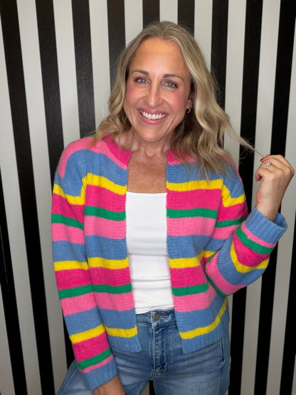 A smiling blonde woman wearing a colorful striped knit cardigan with pink, blue, yellow, and green horizontal stripes over a white tank top and blue jeans, standing against a black and white vertically striped background.