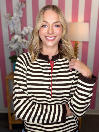A smiling blonde woman wearing a brown and cream striped knit henley sweater with a red button placket, standing in front of a pink and white striped wall.