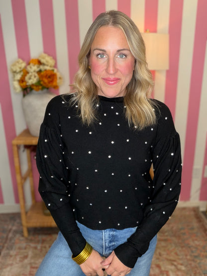 A woman with blonde hair wearing a black long-sleeve mock neck top with white star patterns, paired with blue jeans and gold bracelets, standing in front of a pink and white striped wall.