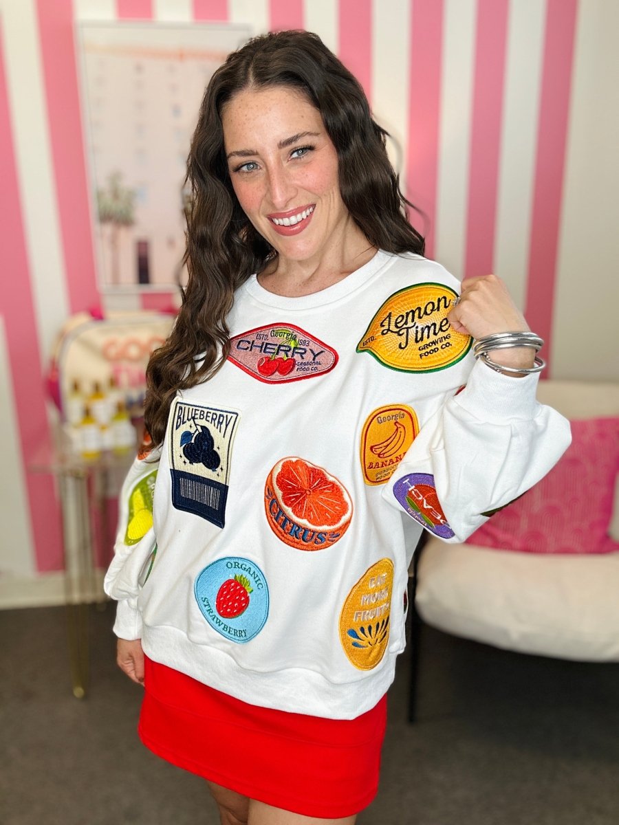 Woman wearing a white sweatshirt with colorful fruit patches in a room with pink and white striped wallpaper.