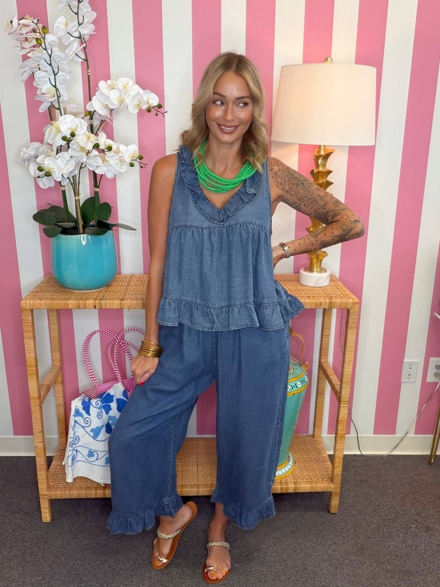A woman modeling a two-piece blue denim set consisting of a ruffled tank top and matching wide-leg pants, styled with a bright green necklace and gold bracelets, standing in front of a pink and white striped wall.