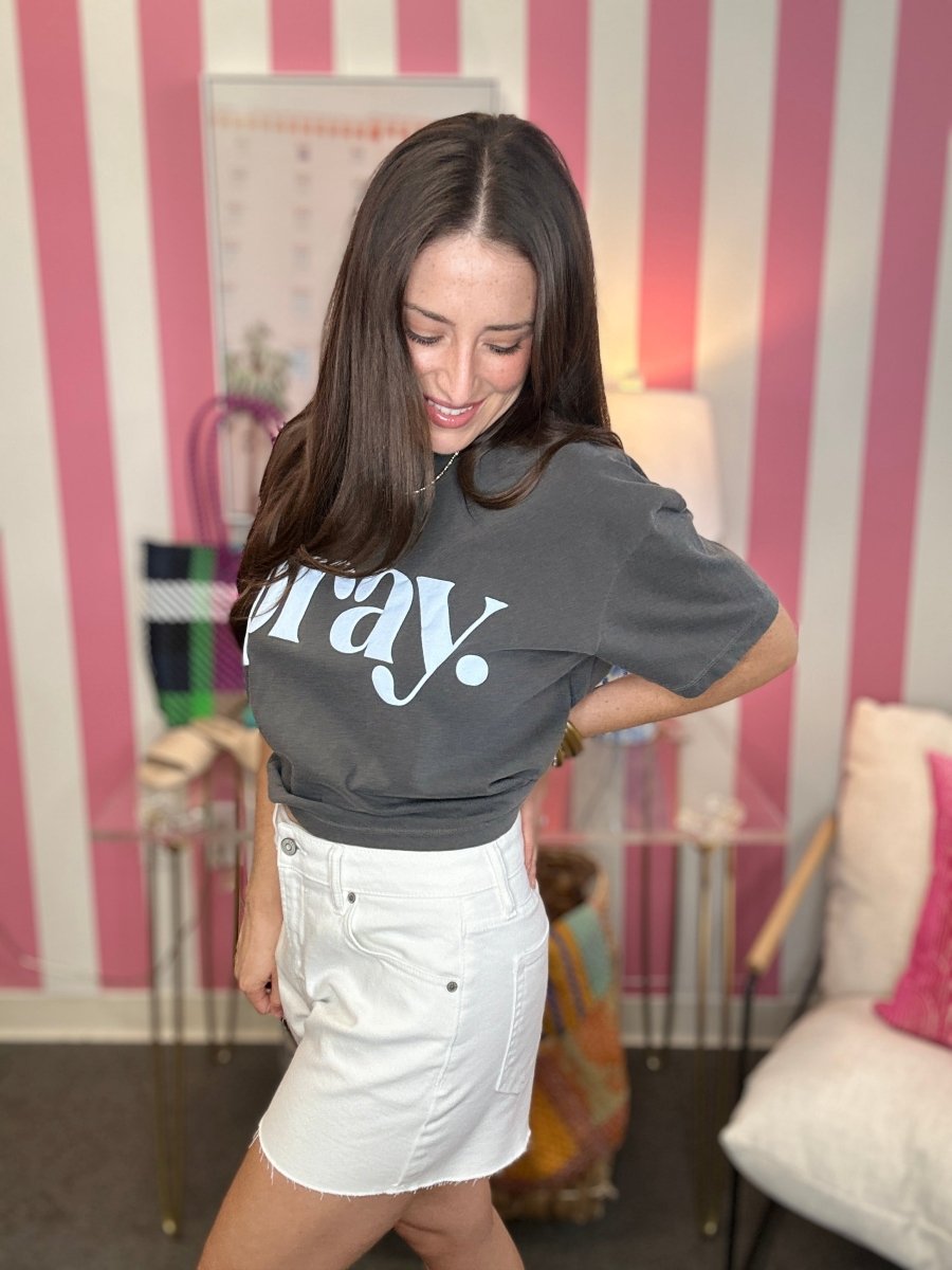 Woman wearing a gray crop top with 'pray' text and white skirt in a room with striped wallpaper.