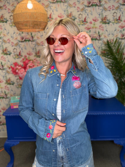 A woman wearing a light blue denim button-up shirt with colorful floral embroidery on the collar, cuffs, and chest pocket, styled with round brown sunglasses and a white top. She is posing in front of a blue bench and a floral wallpaper background.