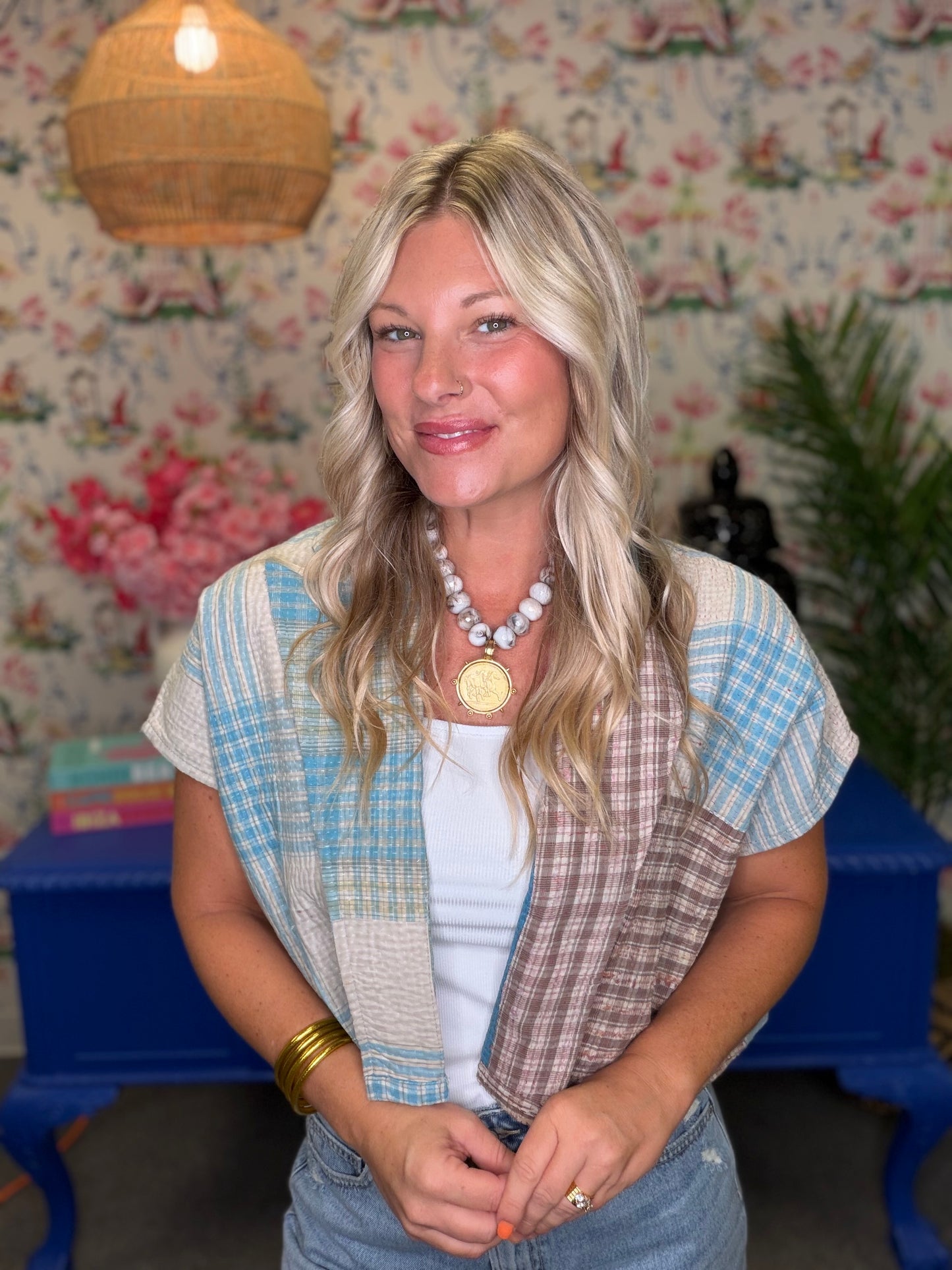 A woman with blonde hair wearing a light blue and brown patchwork vest over a white t-shirt, standing in front of a floral wallpaper background with a blue table.
