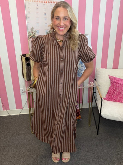 A woman wearing a brown and beige vertically striped midi dress with puff sleeves and a button-down front, standing in front of a pink and white striped wall.