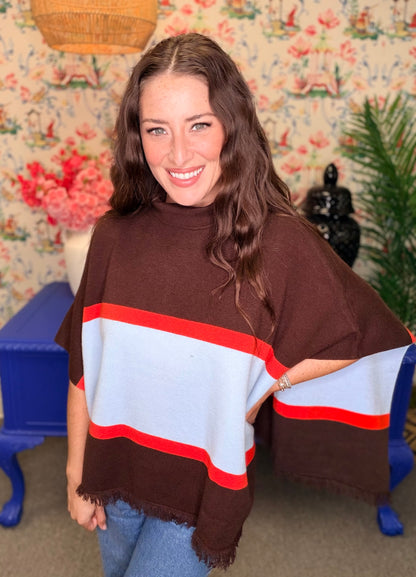 A woman wearing a brown poncho-style top with wide horizontal stripes in light blue and red, standing in a decorated room with floral wallpaper, a blue bench, and a plant.