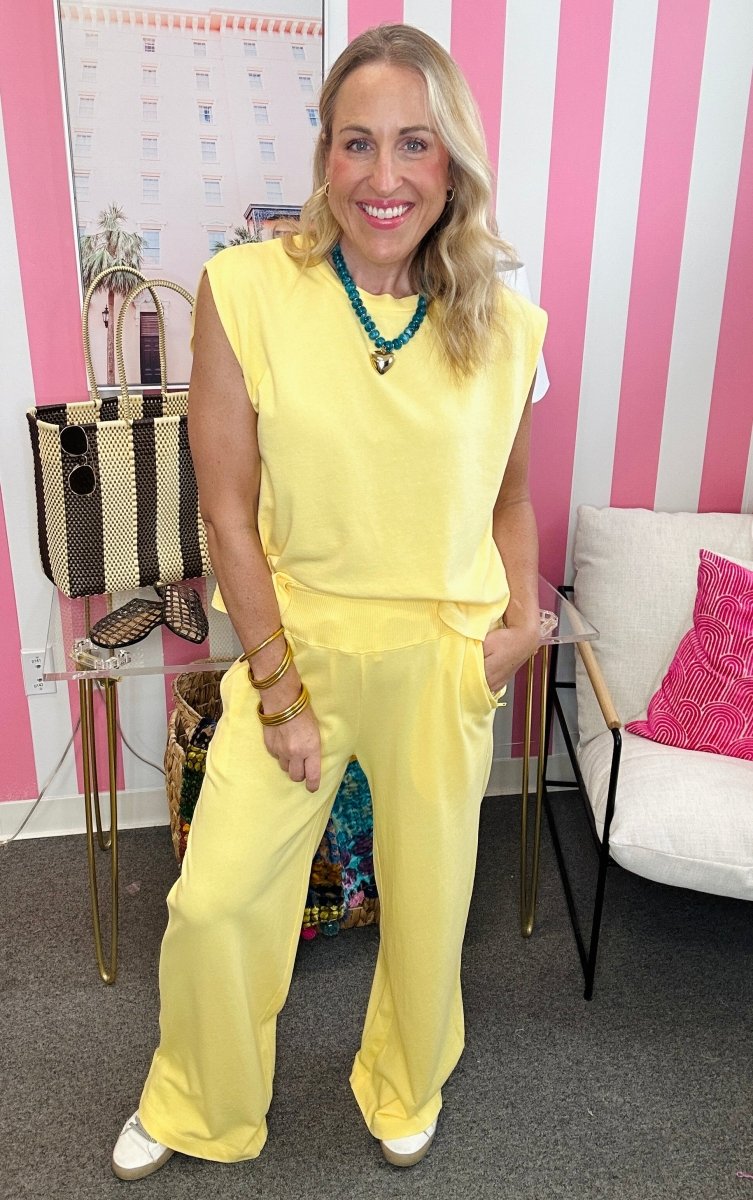 A woman modeling a bright yellow sleeveless jumpsuit with wide legs, standing in front of a pink and white striped wall. She is accessorized with a turquoise necklace and gold bracelets.