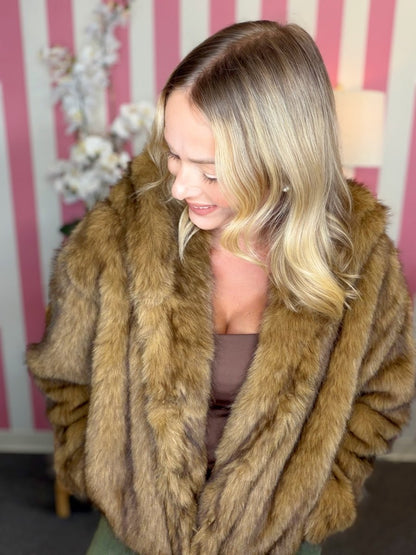 A woman with blonde hair wearing a brown faux fur jacket, looking down and smiling. She is standing in front of a pink and white striped wall with decorative white flowers.