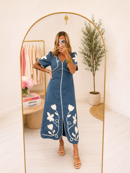A woman takes a mirror selfie in a boutique wearing a blue midi dress with white floral embroidery and white piping. The boutique has a gold-framed arch mirror, a clothing rack with pink and beige shirts, and a potted plant in the background.