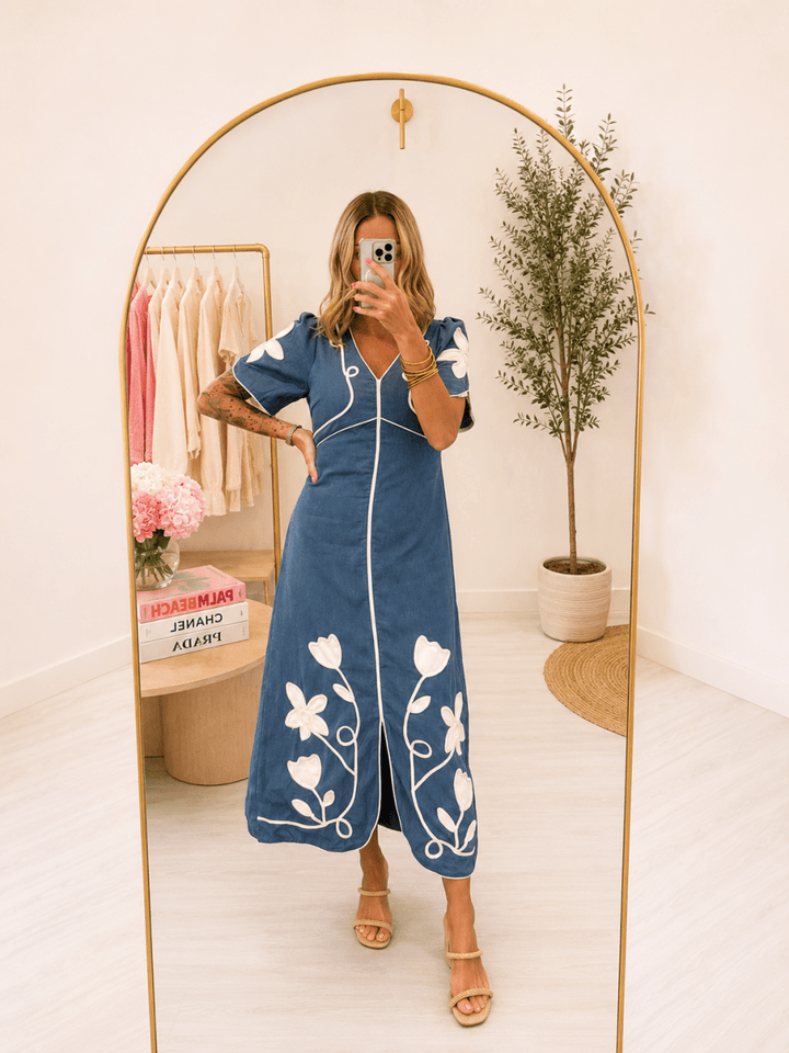 A woman takes a mirror selfie in a boutique wearing a blue midi dress with white floral embroidery and white piping. The boutique has a gold-framed arch mirror, a clothing rack with pink and beige shirts, and a potted plant in the background.