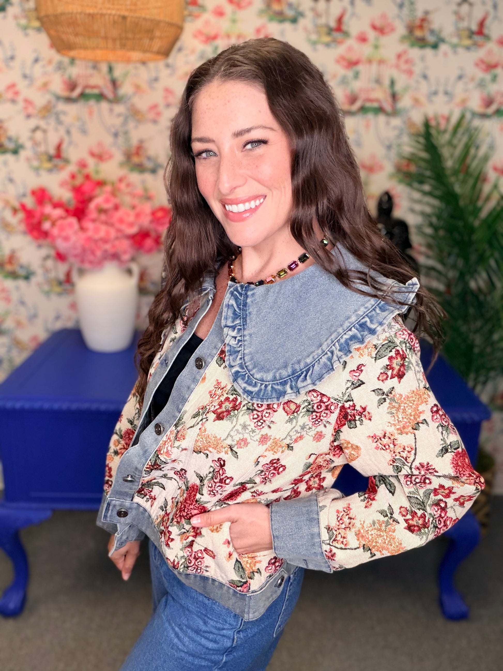 A woman smiling and wearing a cropped denim jacket with a large light blue ruffled collar over a cream floral jacket and blue jeans, standing in front of floral wallpaper and a blue table.
