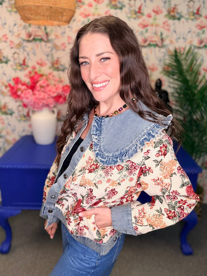 A woman smiling and wearing a cropped denim jacket with a large light blue ruffled collar over a cream floral jacket and blue jeans, standing in front of floral wallpaper and a blue table.