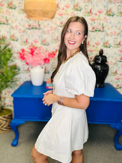 Woman in a white dress standing in a room with floral wallpaper and a blue table.