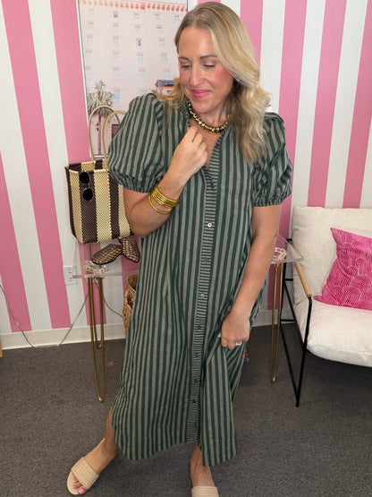 A woman wearing a green and black vertically striped midi shirt dress with puff sleeves and a button-down front, standing in front of a pink and white striped wall.