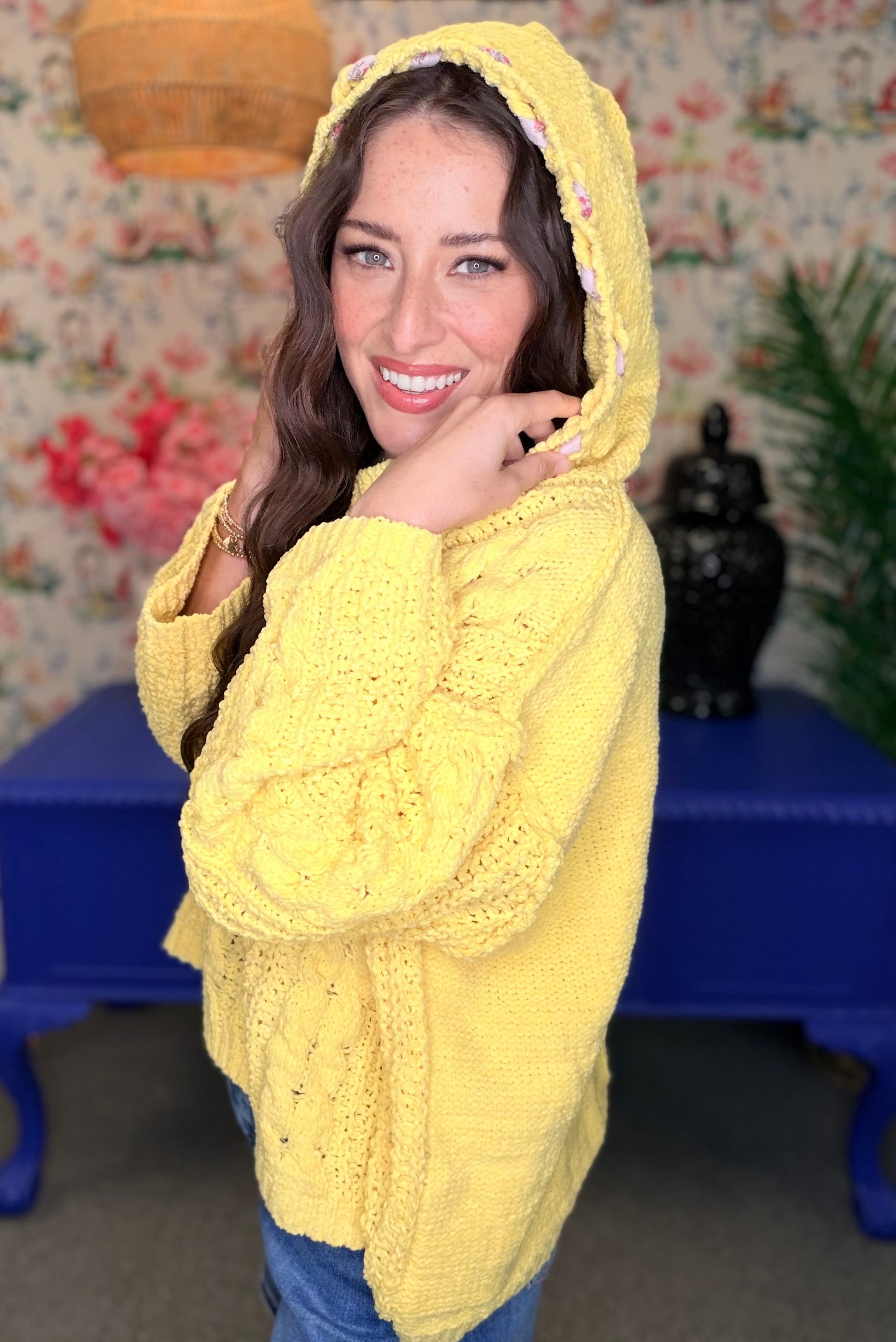 A woman wearing a bright yellow chunky knit hooded sweater, standing in front of a floral wallpaper background with a blue table.