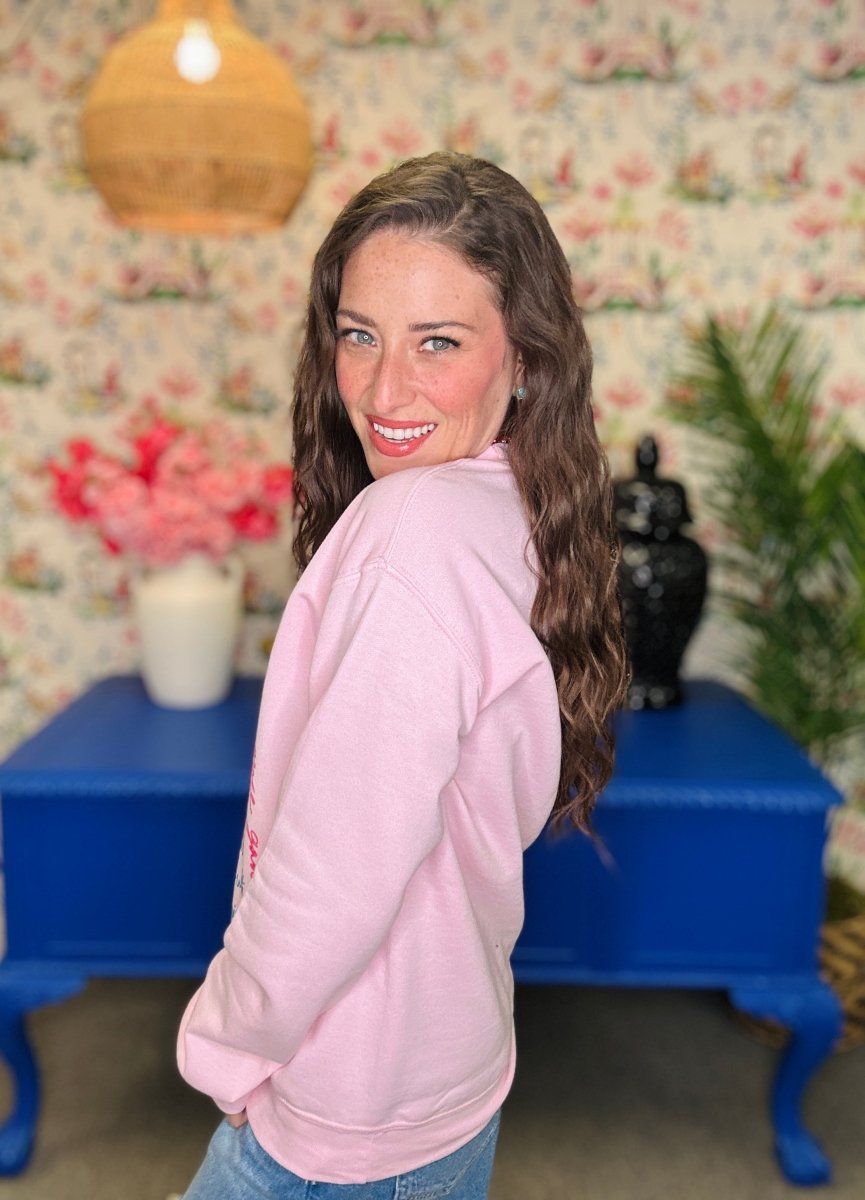 A woman with long brown hair looks back over her shoulder while wearing a light pink sweatshirt and blue jeans, standing in front of a floral wallpaper background with a blue table.