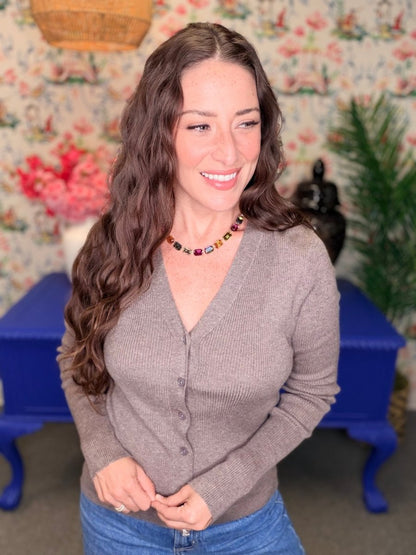 A woman with long wavy brown hair wearing a taupe gray ribbed button-up cardigan and blue jeans, standing in front of a floral wallpaper background with a blue side table.